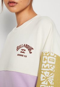 Bluza Billabong z kremowej tkaniny z kolorowymi blokami w odcieniach lawendy i musztardowym. Wyróżniający się biały grafik i logo na musztardowym rękawie.
