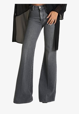 Donna che indossa jeans grigi a vita alta e a gamba larga con un top nero trasparente a maniche lunghe, mano appoggiata sul fianco che mostra anelli e unghie lunghe.