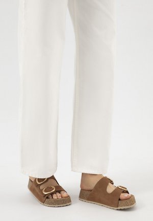 Persoon die witte broek en bruine open-teen slip-on sandalen met twee gespen op kurkzolen draagt, staand op een lichte ondergrond.