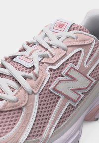Κοντινή λήψη ροζ και λευκού αθλητικού παπουτσιού New Balance με πλεκτό ύφασμα, σουέντ λεπτομέρειες, λευκά κορδόνια και λογότυπο στην πλευρά και στη γλώσσα.