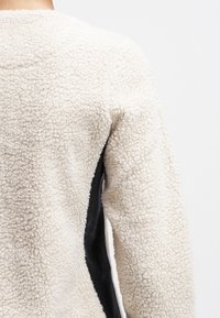 Kremowa kurtka z polaru o teksturowanej powierzchni, z czarnymi bocznymi panelami i okrągłym dekoltem.