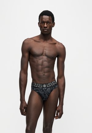 Homme musclé aux cheveux courts portant un slip noir à motif avec une ceinture géométrique, debout sur un fond blanc.