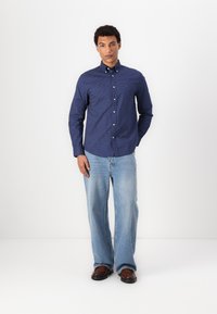 Donkerblauwe overhemd met knopen en witte stippen, lange mouwen en een kraag. Gecombineerd met lichtblauwe wijde jeans en bruine schoenen.