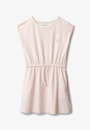 Jeune fille aux cheveux foncés attachés en chignon haut, portant une robe rayée rose et blanche et des baskets beige, debout avec une main dans la poche sur un fond gris.