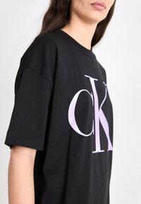 Czarna bawełniana koszulka z dużym białym i lawendowym logo "CK" z przodu. Krótkie rękawy i luźny krój.