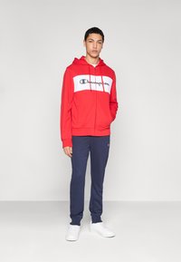 Κόκκινο φούτερ με κουκούλα και λευκή πινακίδα στο στήθος που εμφανίζει το λογότυπο "Champion", σε συνδυασμό με μπλε σκούρο παντελόνι φόρμας και λευκά αθλητικά παπούτσια.