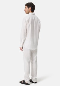 Weißes Leinenhemd und -hose, lockere Passform, lange Ärmel mit Knopfmanschetten. Getragen mit schwarzen Sandalen. Einfache, glatte Textur, neutrale Farbe.