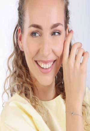 Bracelet en argent en forme de cœur au poignet, avec une chaîne délicate. Le modèle a des cheveux bouclés et porte un haut jaune. L'arrière-plan est blanc uni.