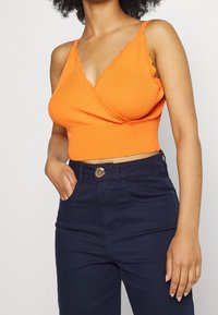 Oranžový ribkovaný cropped top se zvlněn�ými okraji a úzkými ramínky, v kombinaci s tmavě modrými kalhotami s kulatým knoflíkem a kapsami.