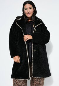 Schwarzer Fleece-Mantel mit weicher Textur, großem Kragen, kontrastierenden cremefarbenen Nähten, zwei Knöpfen und einer lockeren Passform. Getragen über Tiermotiv-Hosen.