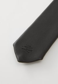 Schwarze Ledernocke mit strukturiertem Oberfläche, spitzem Ende und einem dezenten, geprägten Logo nahe der Spitze. Glatte Oberfläche.