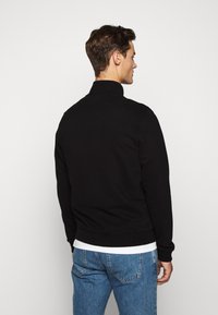 Svart sweatshirt med hög krage, ribbade mudd och nederkant, tillverkad av ett mjukt material. Buren över en vit t-shirt och kombinerad med blå jeans.