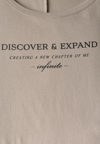 Ljusbeige t-shirt med svart text: "UPPTÄCK & UTVIDGA, SKAPA ETT NYTT KAPITEL AV MIG oändligt." Mjuk bomullstyg, rund halsringning.