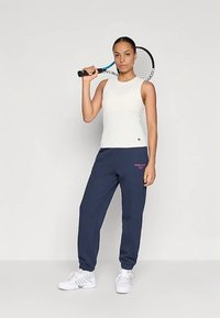 Γυναίκα σε λευκή αμάνικη μπλούζα, ναυτικές φόρμες και λευκά αθλητικά παπούτσια, κρατώντας ένα ρακέτα τένις που έχει ακουμπήσει στον ώμο της, στέκεται μπροστά σε απλό φόντο.