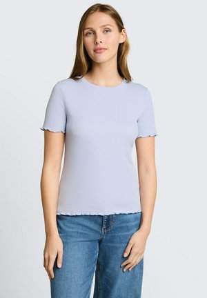 Jeune femme aux longs cheveux blonds portant un haut bleu clair ajusté à manches courtes et un jean bleu, debout devant un fond uni.