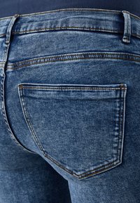 Los jeans de mezclilla azul presentan una textura suave con un desvanecimiento sutil. La costura es visible en hilo de contraste y hay dos bolsillos traseros.