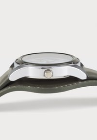 Stainless steel klokke med sølvkasse, svart skive og en teksturert sølvkrone; har en grønn lærrem.