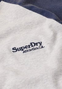 Zbliżenie szarej tkaniny z ciemnoniebieskim haftowanym logo "SuperDry Athletics Co." oraz ciemnoniebieskim brzegiem tkaniny u góry.