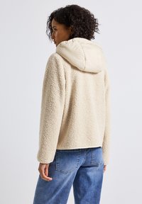 Femme portant une veste en polaire crème avec une capuche rembourrée et un jean bleu, tournée sur le côté devant un fond blanc uni.
