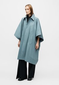 Lichtblauwe oversized jas met kraag, korte mouwen en een voorzak. Gedragen over zwarte broek met wijd uitlopende pijpen en schoenen. Gladde textuur.