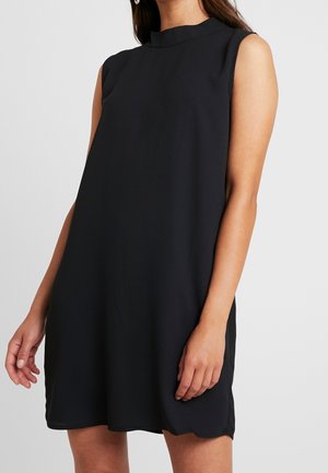 Femme portant une robe noire sans manches à col montant, vue des épaules jusqu'à mi-cuisse sur un fond uni.