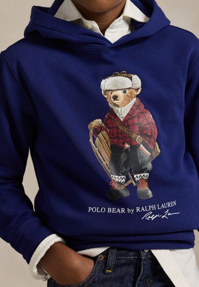 Sweatshirt azul-marinho com capuz, apresentando um gráfico de um urso vestido para o inverno, segurando um trenó. Inclui padrão xadrez, detalhes em branco e logótipo bordado.