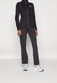 Svart fleece zip-up-jacka och matchande byxor, tillsammans med grå löparskor. Funktioner inkluderar fickor med dragkedja och logodetaljer.