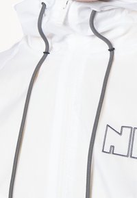 Bílá bunda se zipem na přední straně, s vysokým límcem, šedými šňůrkami a černým vyšitým logem. Hladká, lehká látka.