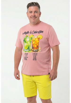 Mężczyzna w różowej koszulce z ilustracjami koktajli mojito i cuba libre oraz żółtych szortach, stojący na białym tle.