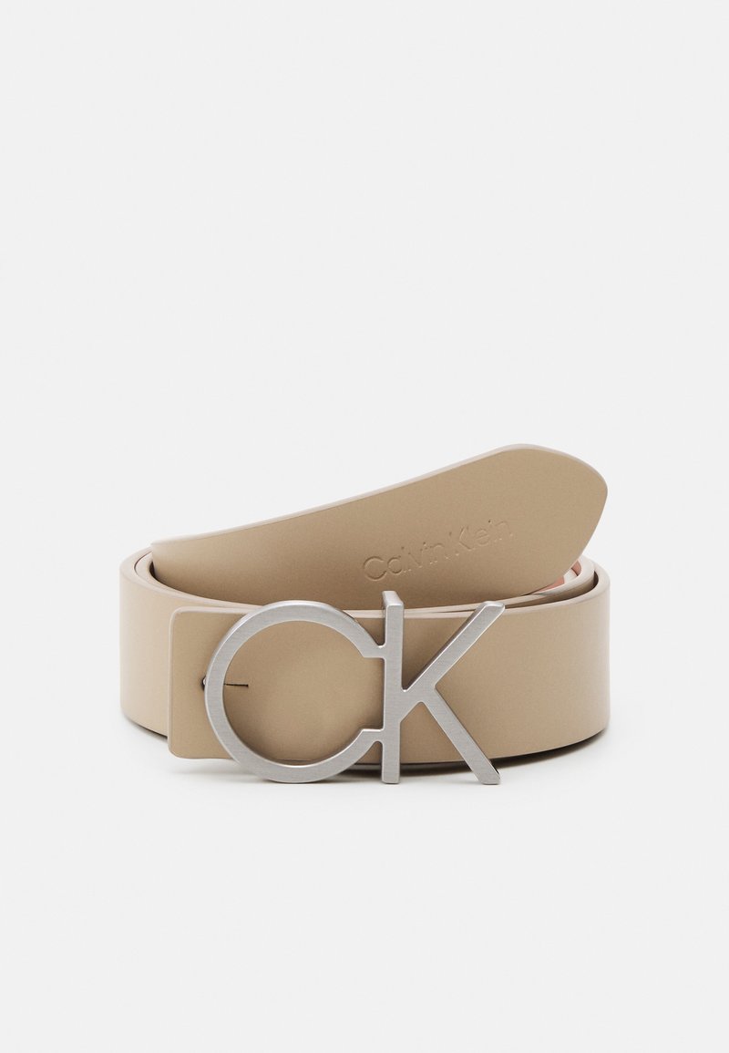 Cinturón de cuero beige con una hebilla metálica redonda en tono plata que presenta un diseño de "CK". El lado interior tiene el logotipo en relieve de "Calvin Klein".