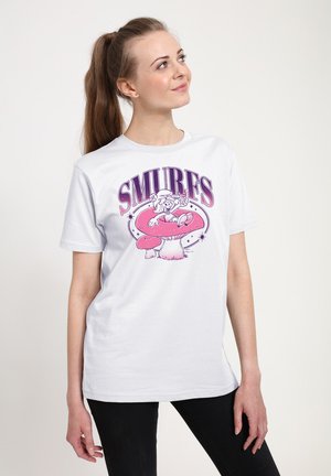Jonge vrouw met een paardenstaart die een wit Smurfen-thema t-shirt draagt met een roze paddenstoel en figuurafbeelding, kijkt iets omhoog en naar rechts.