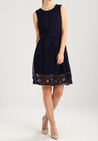 Námořnické modré šaty bez rukávů vyrobené z texturované látky, s kruhovými výřezy na lemu a přiléhavým pasem.