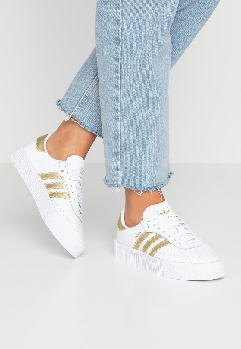 Persona che indossa sneakers bianche con strisce dorate e dettagli sul tallone, abbinate a jeans azzurri con orlo sfrangiato, in piedi su una superficie bianca.