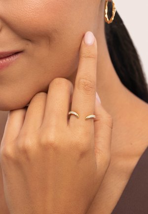 Anillos de oro con diseños curvos, uno adornado con pequeños cristales brillantes. La mano del modelo muestra los anillos sobre una piel suave.