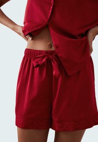 Red satin pajama shorts with an elastic waistband and bow tie detail, featuring lace trim at the hem for added texture and elegance.