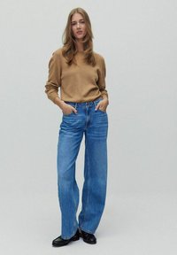 Pull en maille marron avec une coupe décontractée, associé à un jean en denim bleu à jambes larges. Le modèle se tient les mains dans les poches devant un fond gris clair.