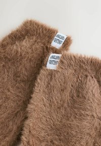 Braune Kunstpelz-Beinwärmer mit einer sanften Textur, versehen mit weißen Etiketten, die mit einem dekorativen schwarzen Muster verziert sind.