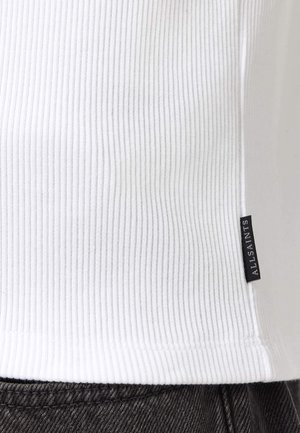 Top in maglia a coste bianca con una piccola etichetta di tessuto nera con la scritta "ALLSAINTS" infilata in pantaloni di jeans grigio scuro.