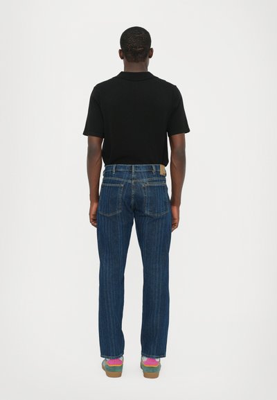 Jeanși din denim de culoare închisă, cu croială dreaptă, având două buzunare la spate și un patch din piele maro. Cămașă polo neagră și adidași colorați.