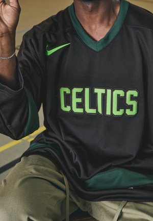 Person wearing dark green and black Celtics sports jersey with green Nike logo, seated with one arm raised wearing a silver bracelet.