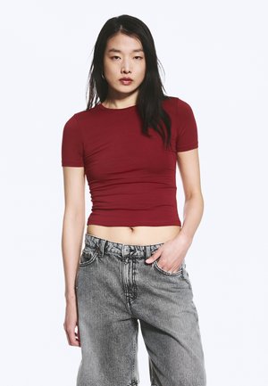 Frau mit langen dunklen Haaren, die ein eng anliegendes dunkelrotes kurzes T-Shirt und lockere graue Jeans trägt, steht mit einer Hand in der Tasche.