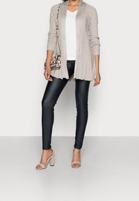 Cardigan côtelé beige sur un t-shirt à col en V blanc, associé à des leggings imitation cuir noirs et des sandales à talons carrés nude. Sac à épaule à motifs.