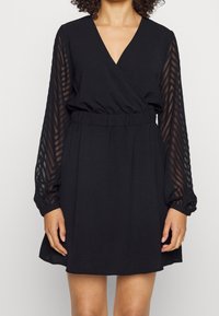 Femme portant une robe noire à décolleté en V et à manches longues transparentes présentant des motifs en chevrons, se tenant devant un fond uni.