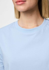 T-shirt de algodão azul claro com decote redondo. O tecido parece suave e liso, apresentando uma textura sutil e uniforme.