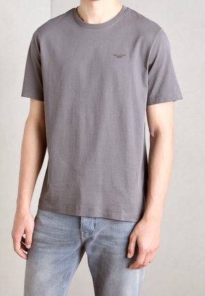 Mężczyzna noszący gładki szary t-shirt z krótkim rękawem z małym logo "Marc O'Polo DENIM" na piersi oraz jasnoniebieskie dżinsy.