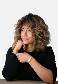 Bracciale d'argento con anelli circolari e ovali, indossato su un braccio. Maglietta a maniche lunghe nera con scollatura a scallop. Capelli ricci incorniciano il viso.
