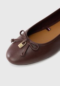 Barna bőr balettcipő, lekerekített orral, csavart masni díszítéssel és egy kis arany lakat részlettel. Simaság és minimalista dizájn jellemzi.