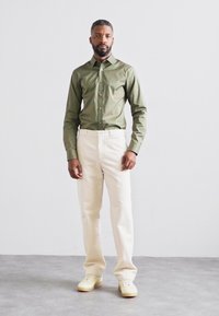 Cămașă verde măsliniu cu nasturi, cu mâneci lungi, asociată cu pantaloni drepți de culoare crem și adidași bej deschis. Design simplu.