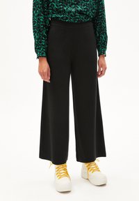 Zwarte wijde broek met een elastische tailleband, gemaakt van zacht materiaal. Gecombineerd met witte sneakers met gele veters.