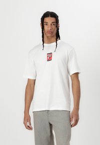 Hvid t-shirt med korte ærmer, der har et centralt rødt 7UP-logo på en sort baggrund. Stoffet ser blødt ud; tøjet har en afslappet pasform.